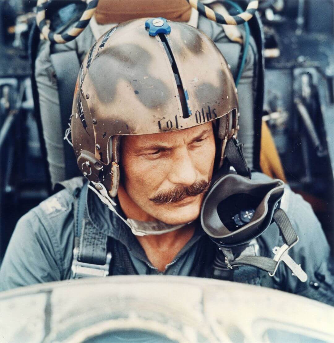 A man with a mustache wearing flight gear sits in the cockpit of a fight plane.